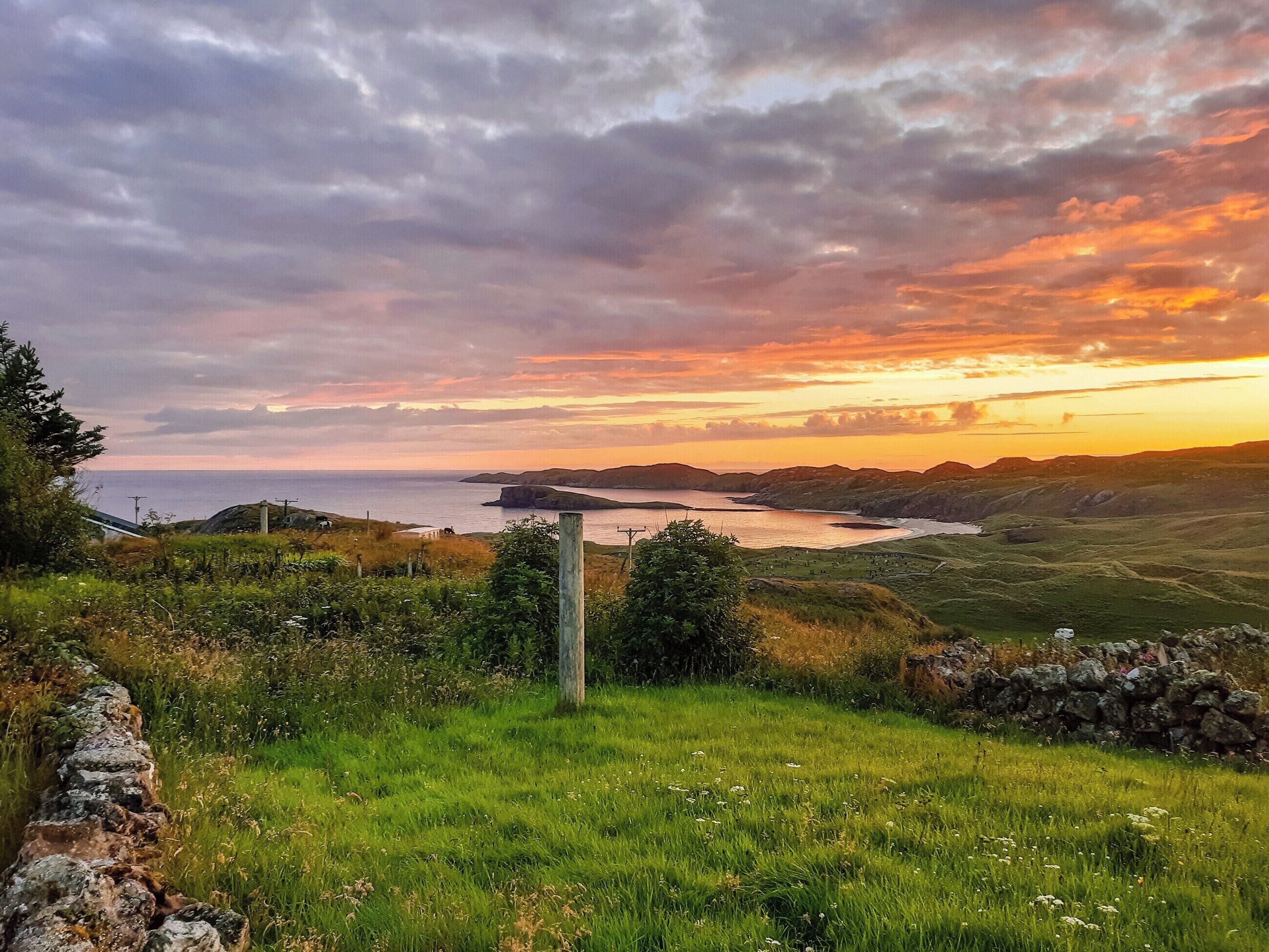 A coastal landscape with a grassy foreground, a stone wall, and a view of the sea and hills during sunset.