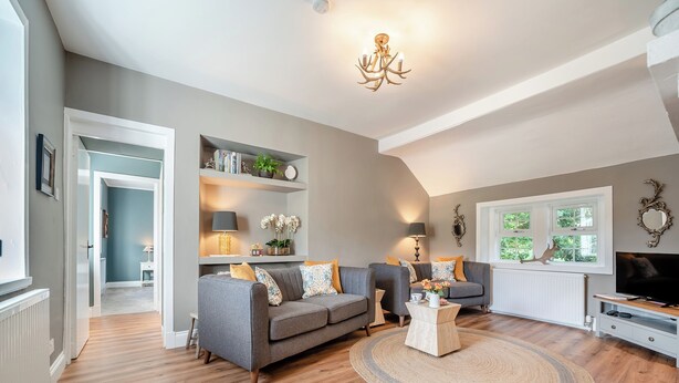 Een woonkamer met een grijze bank, een salontafel en een televisie. Er zijn planken met decoratieve objecten en een raam met uitzicht op groen.