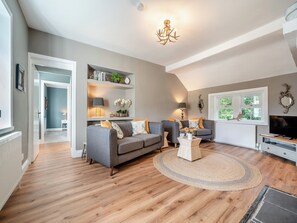 Un salon avec un canapé gris, une table basse et un téléviseur. On y trouve des étagères ornées d’objets décoratifs et une fenêtre donnant sur un paysage verdoyant.