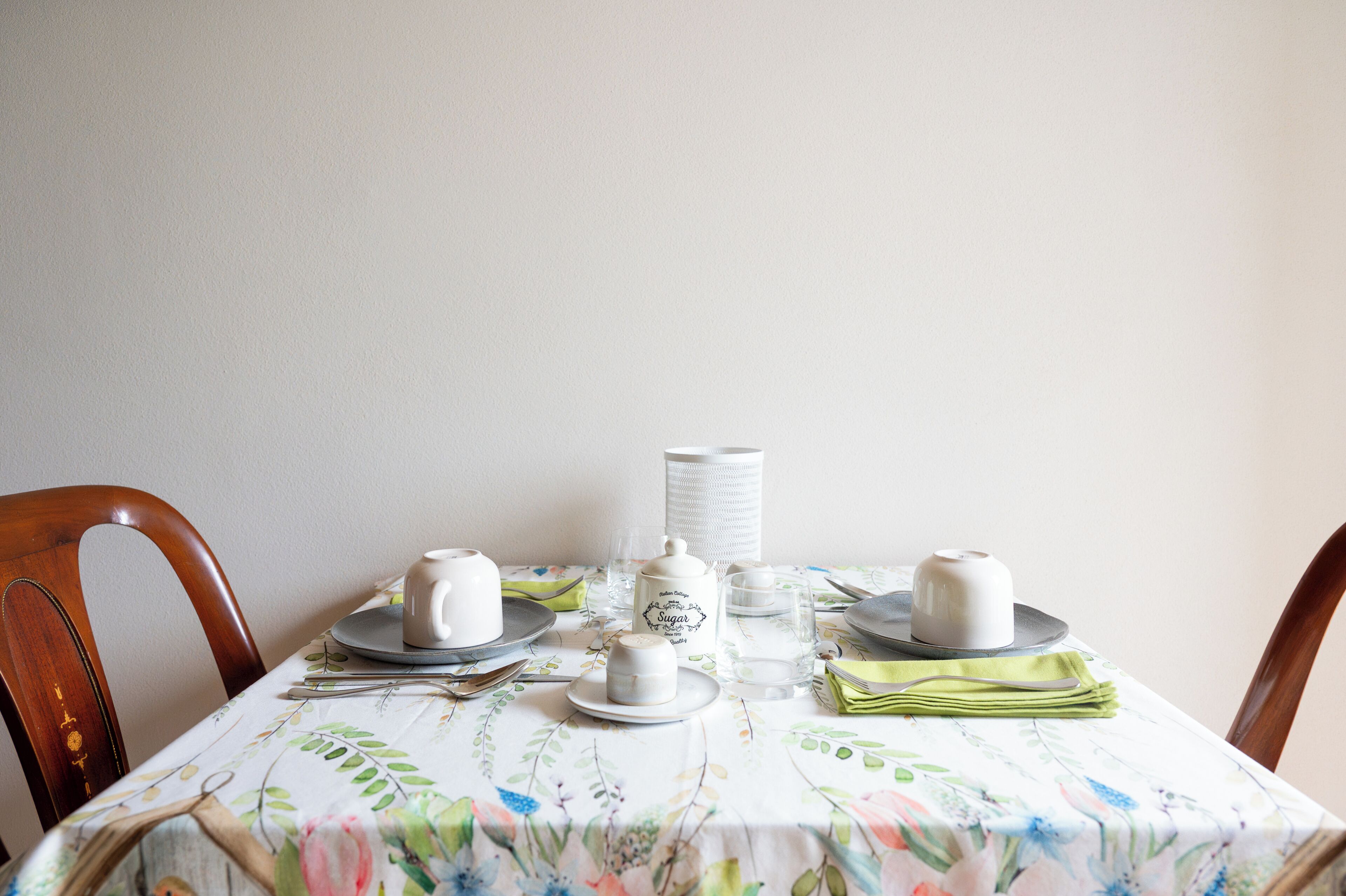 Una mesa de comedor con teteras blancas, recipientes de azúcar y cristalería sobre un mantel estampado con motivos florales.