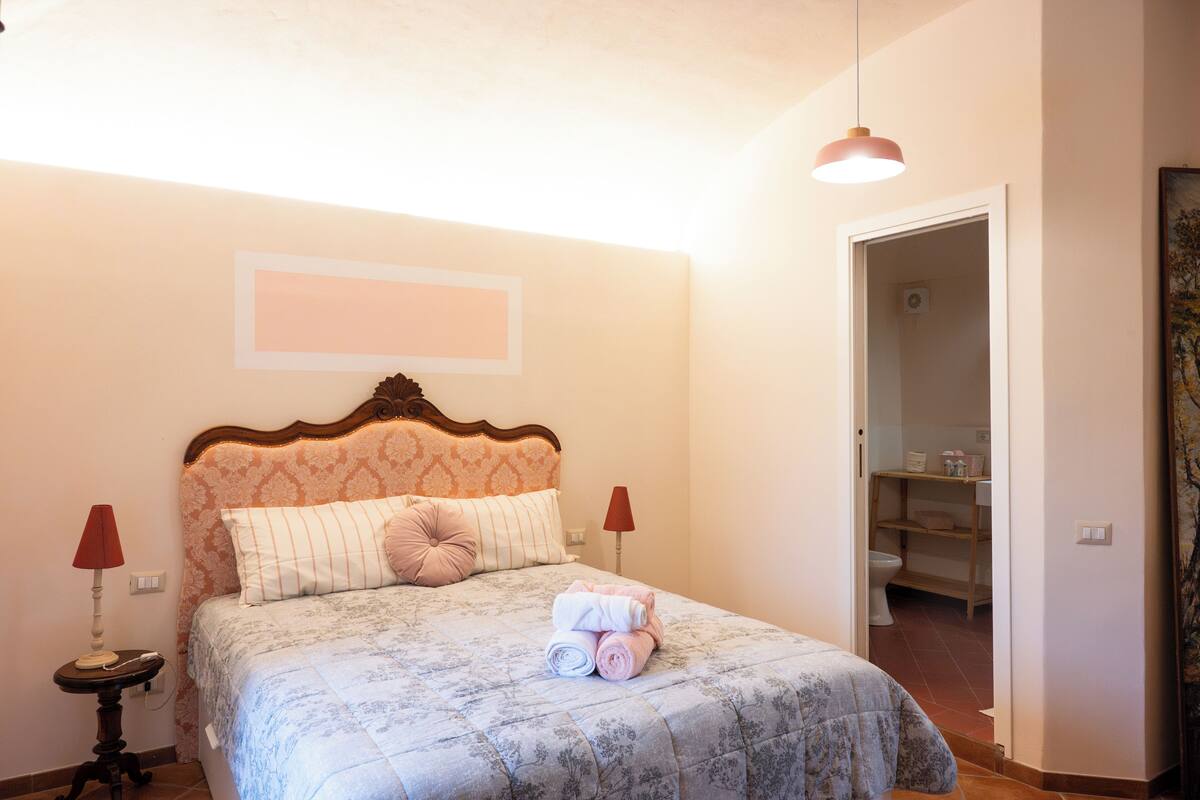 A bedroom with a bed, two bedside lamps, a bedside table, and a door leading to a bathroom.