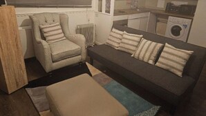 Un salon avec un canapé, un fauteuil et un petit pouf. On aperçoit un radiateur et un lave-linge au fond de la pièce.