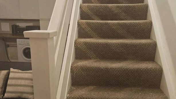 Un escalier recouvert de moquette, avec une rampe blanche, et une machine à laver en arrière-plan.