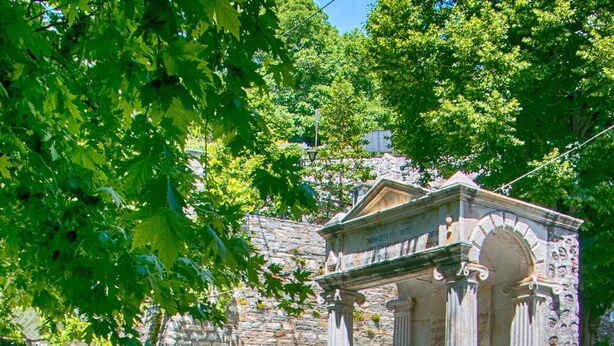 Un espace extérieur aménagé avec des tables recouvertes de nappes à carreaux et des chaises en bois, une structure en pierre ornée de colonnes, et un escalier menant à un bâtiment.