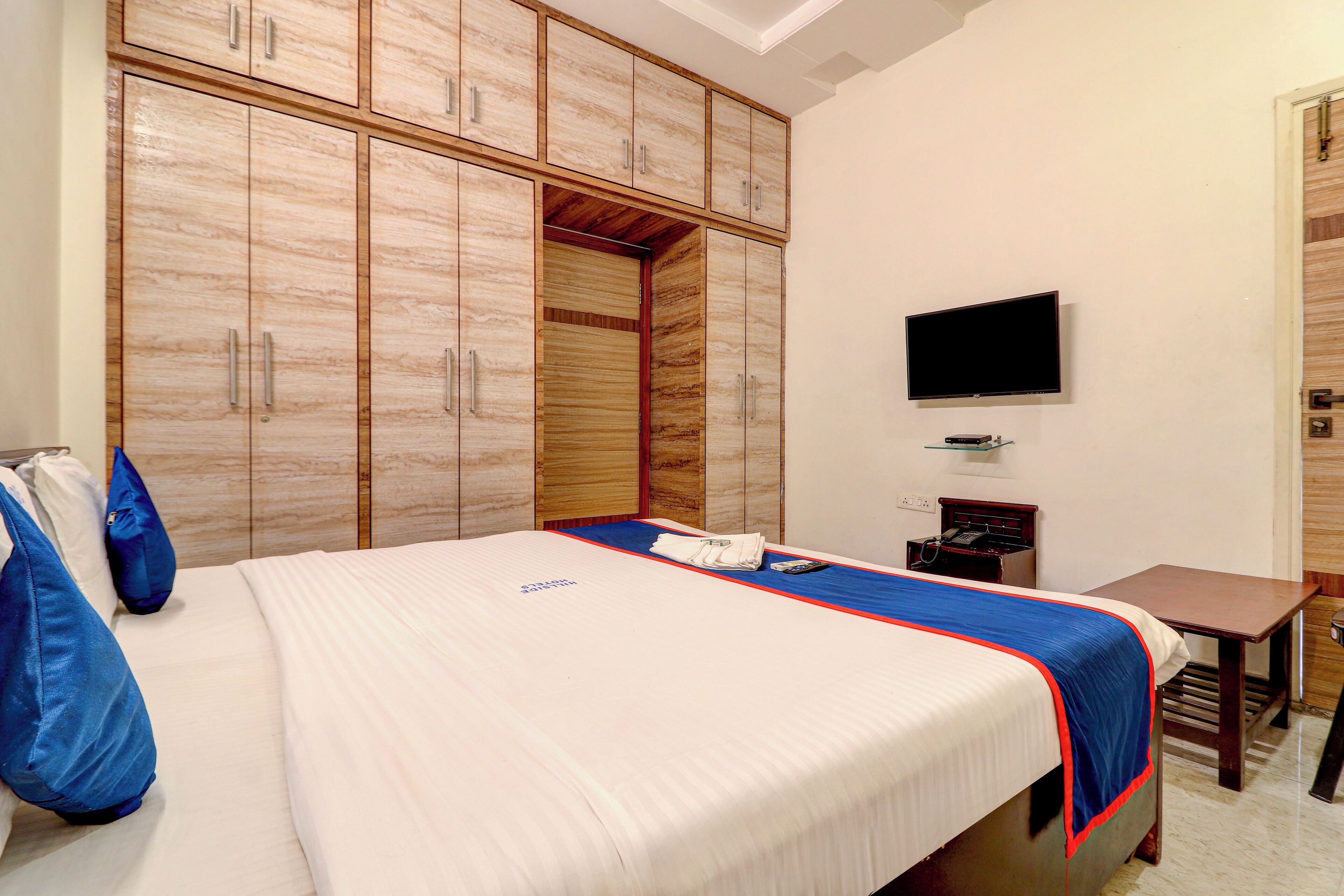 A hotel room with a large wardrobe, a bed with a blue and white bedspread, a flat-screen TV mounted on the wall, and a small wooden table.