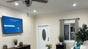 A modern living room with a flat-screen TV, ceiling fan, dark wooden furniture, and a large window with curtains.
