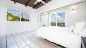 Un dormitorio con cama, ventilador de techo y ventanas amplias con vista a follaje.