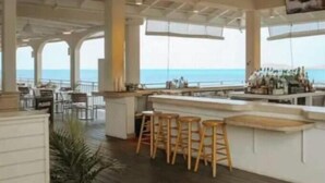 Una zona de bar con taburetes de madera, una barra y vista al mar.