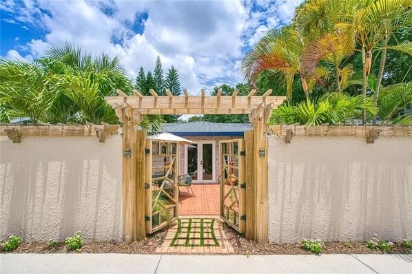A wooden pergola entrance to a property with a paved patio and a view of lush greenery.