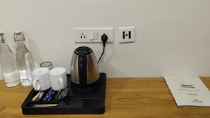 Un coin café avec une bouilloire, des tasses et des bouteilles sur une table en bois, une prise électrique et un menu affiché au mur.