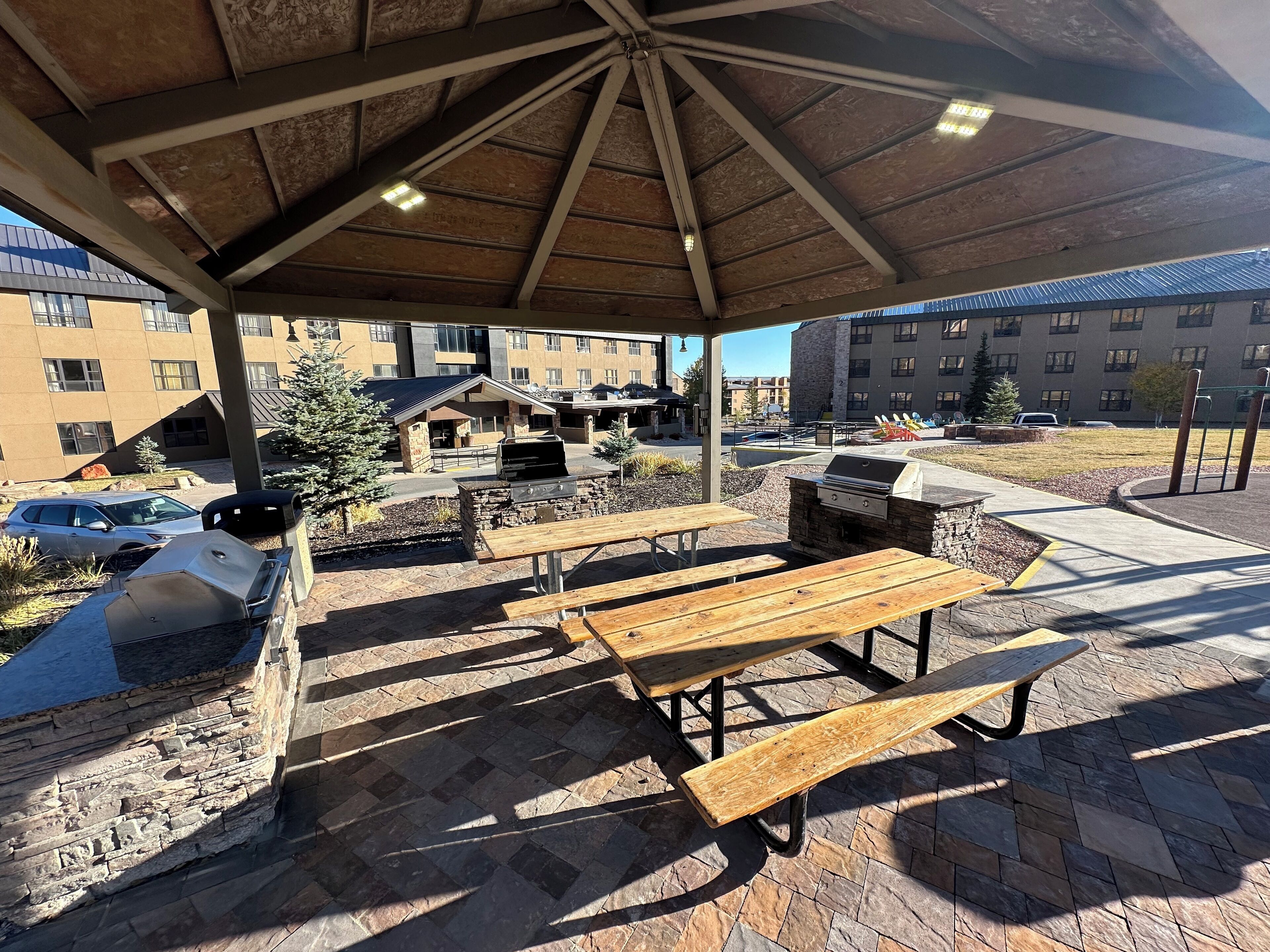 Une terrasse extérieure avec des tables de pique-nique et un barbecue, entourée de murs en pierre et abritée par une tonnelle en bois.