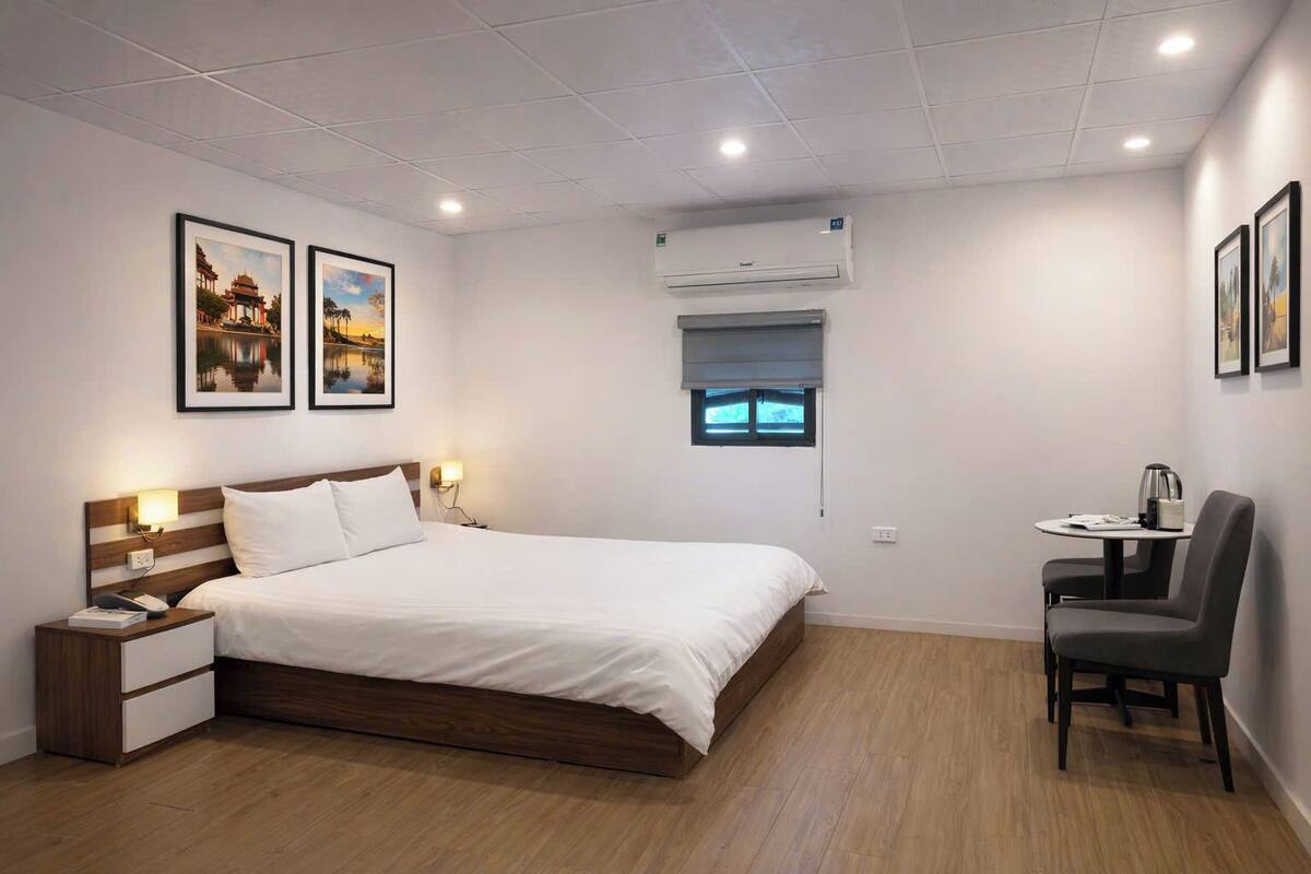 A hotel room with a bed, bedside table, lamp, air conditioning unit, window, and a small table with a chair.
