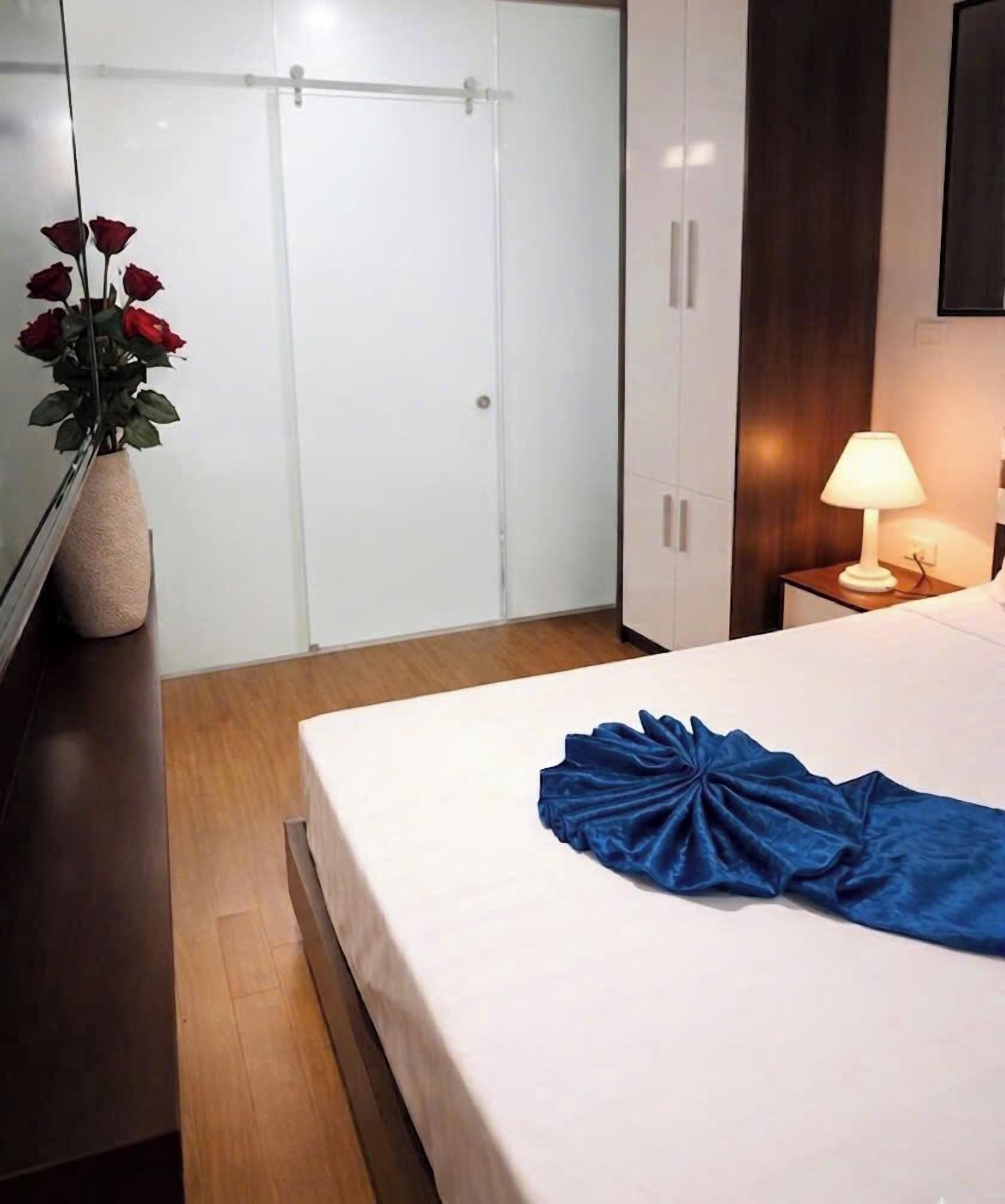 A hotel room with a bed, a nightstand with a lamp, a vase with flowers, and a wardrobe.
