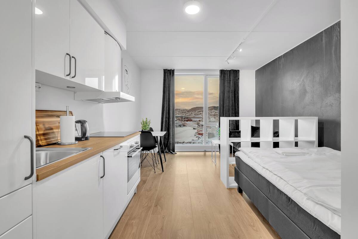 A modern hotel room with a kitchenette, a bed, and a view of a cityscape.