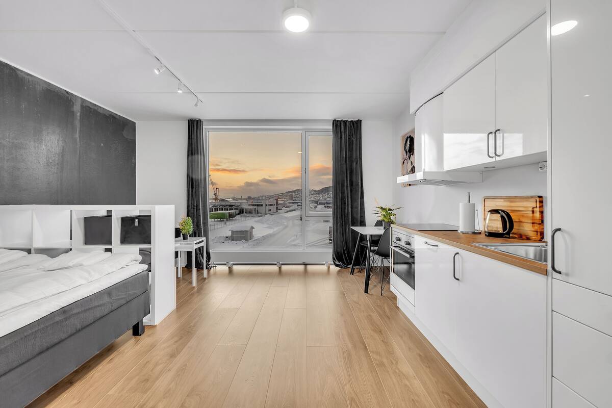 A modern hotel room with a bed, desk, and kitchenette.