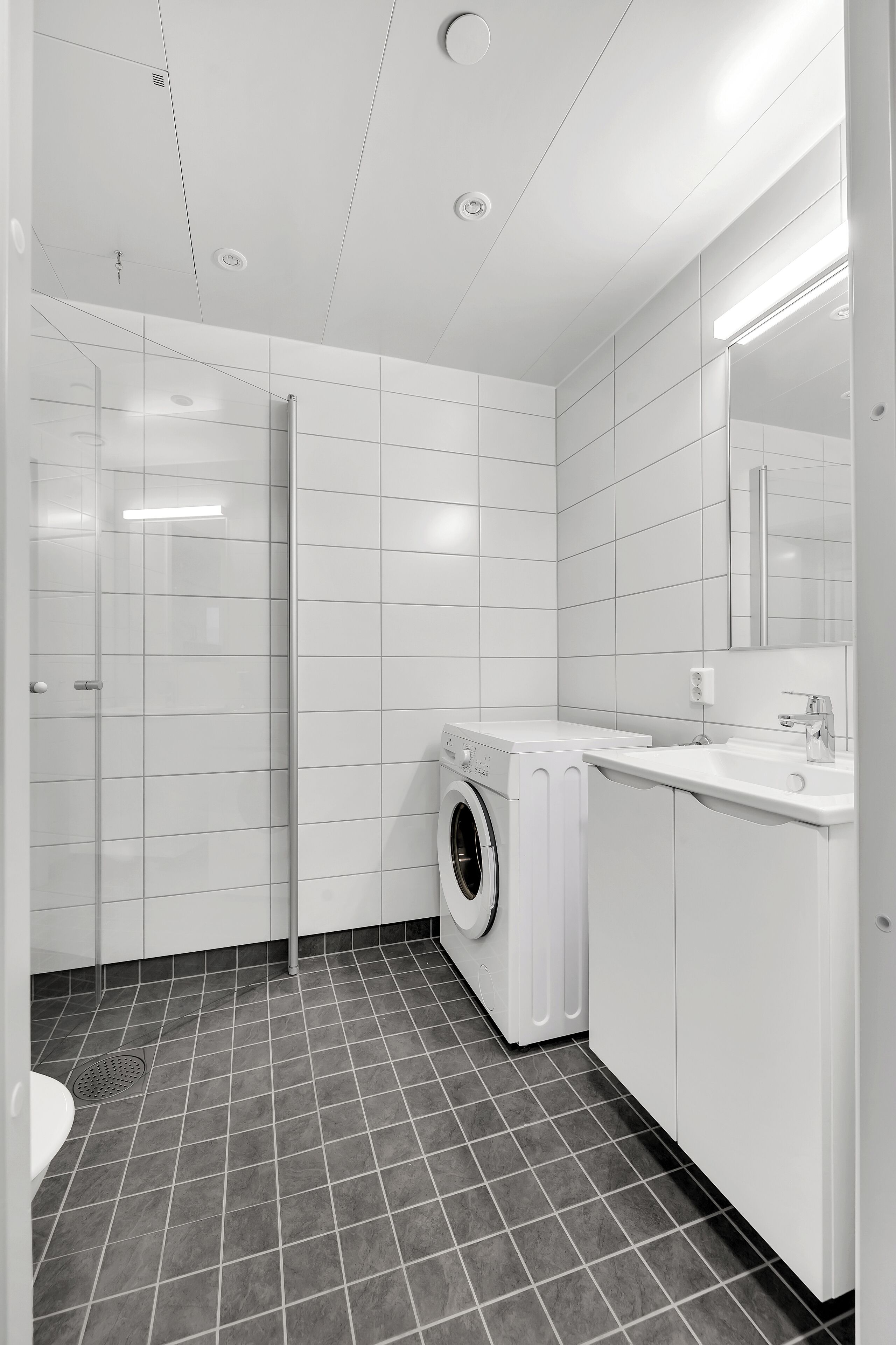 A modern bathroom with a glass shower enclosure, a built-in washing machine, and a white sink.