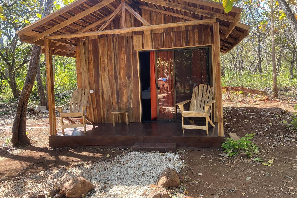 A rustic wooden cabin with a porch, two wooden chairs, and a gravel path leading to the entrance.