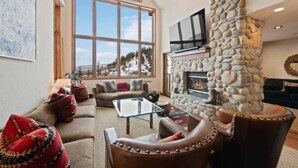 A cozy living room with a stone fireplace, large windows offering a view of snowy mountains, and comfortable seating. - Highlands Lodge 401 - 3 Bedroom Residence (Beaver Creek)