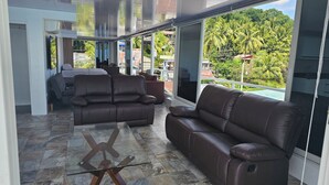 A modern living room with leather sofas, a glass coffee table, and large sliding glass doors leading to a balcony with a view of palm trees and buildings. - loft rooftop beachfront with an amazing view (Island Garden City of Samal)