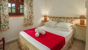 Ein ordentlich bezogenes Bett mit roter Decke und weißen Kissen, zwei Nachttischlampen, ein Fenster mit floralem Vorhang und ein Holznachttisch.