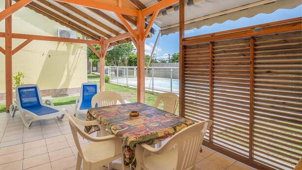 Eine überdachte Terrasse mit Tisch und Stühlen, ein Pool im Hintergrund und ein Holzzaun.
