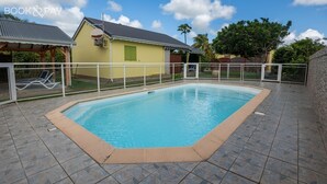 A swimming pool with a tiled deck, a covered seating area, and a building with a green window in the background. - Villa Muscade - Maison avec piscine partagée (Saint-Francois)
