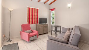 A living room with a pink chair, grey sofa, and a TV on a cabinet. - Villa Muscade - Maison avec piscine partagée (Saint-Francois)