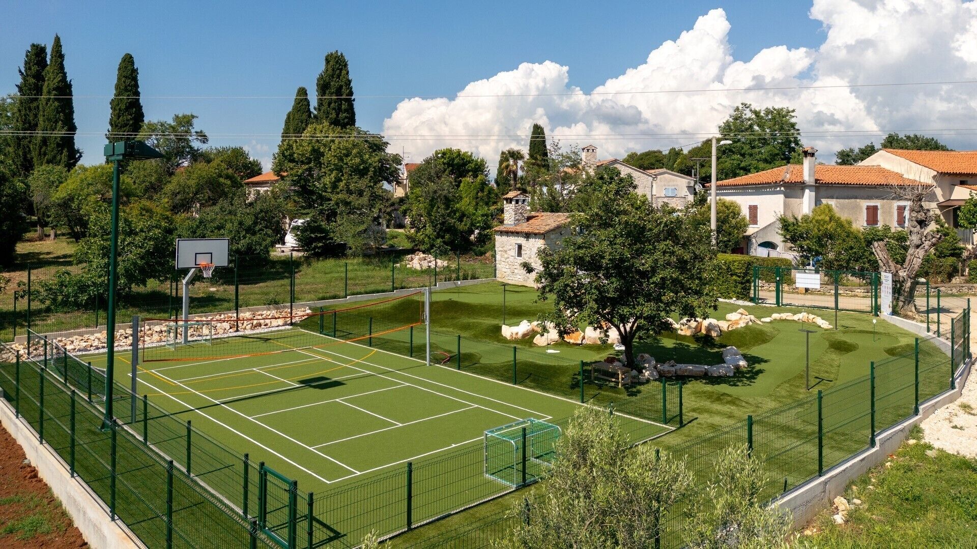 A tennis court surrounded by a green fence, with stone buildings and trees in the background.