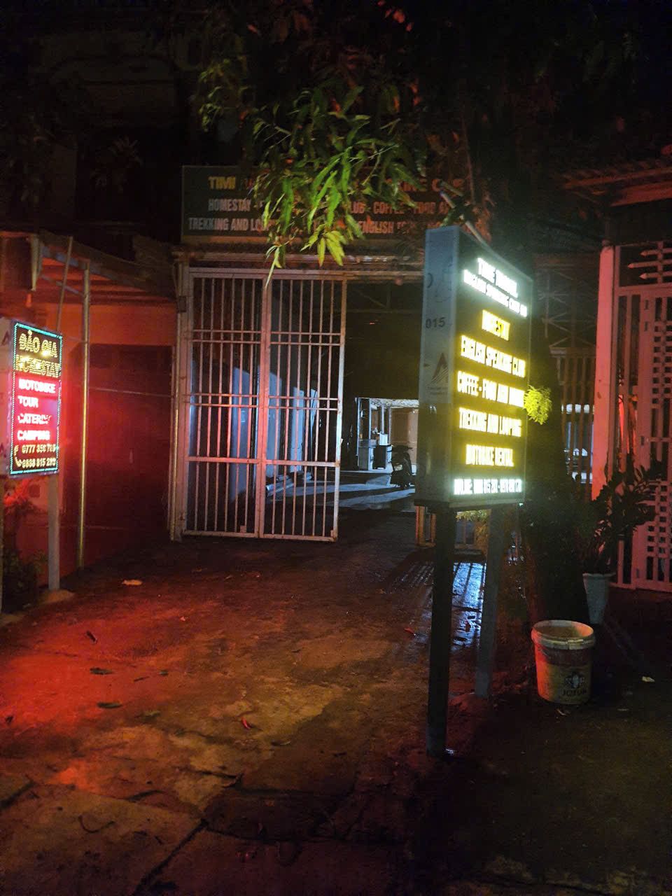 A nighttime view of a building with a gated entrance, a neon signboard, and a potted plant.