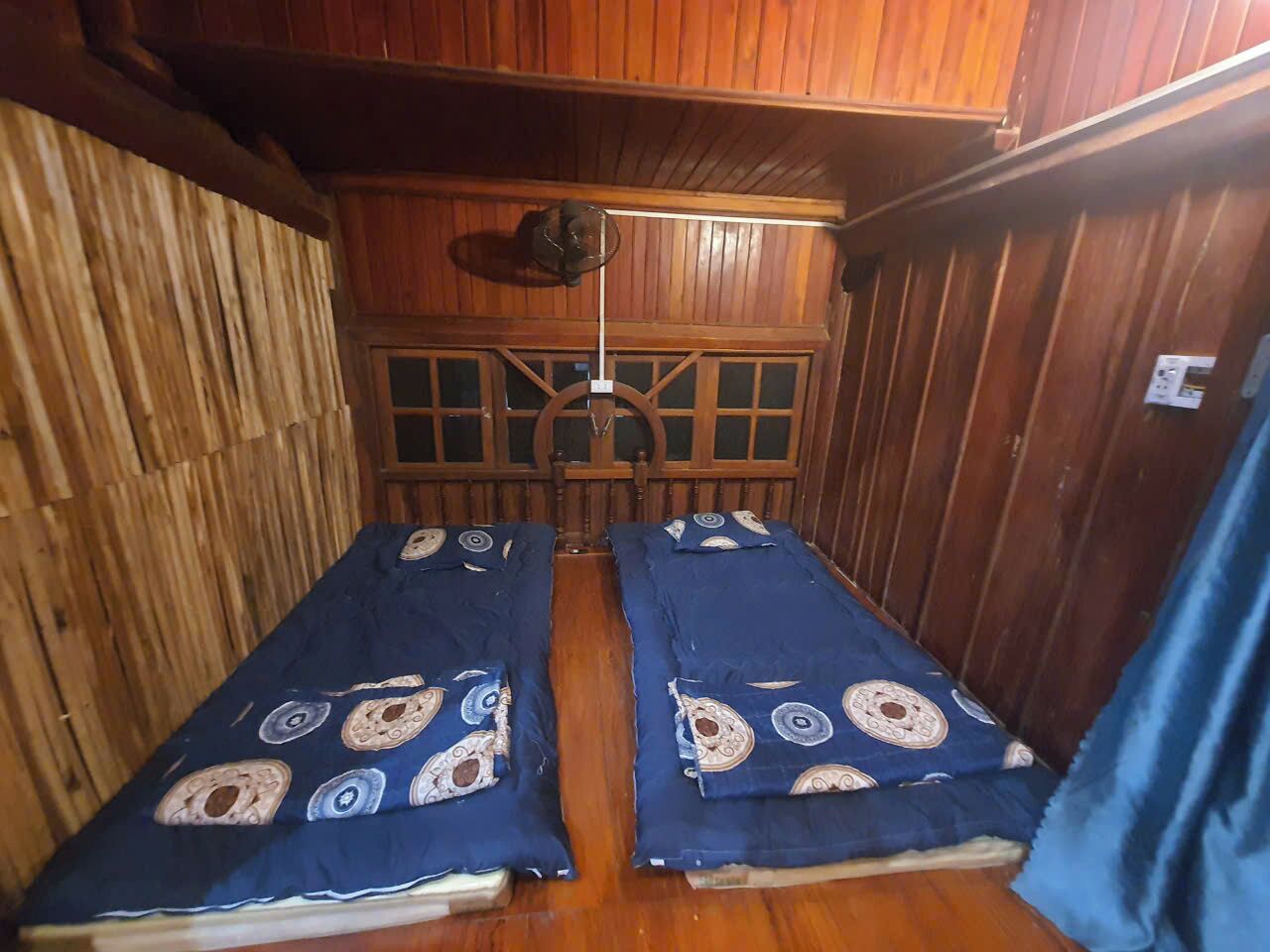 A room with wooden walls and a ceiling fan. There are two beds with blue bedsheets and patterned pillows.