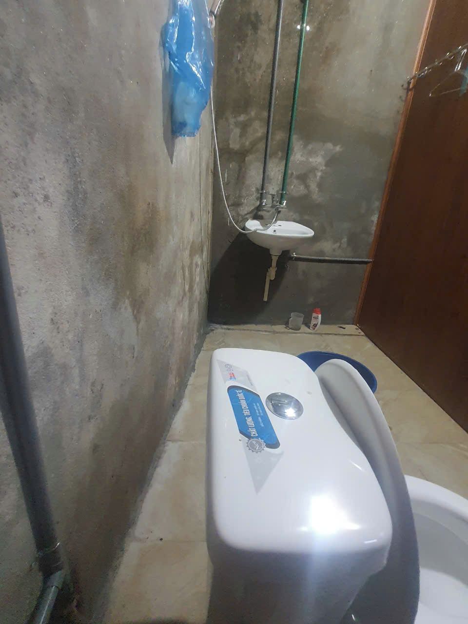 A small, dimly lit bathroom with a white toilet, a blue plastic bag hanging on the wall, and a green pipe.