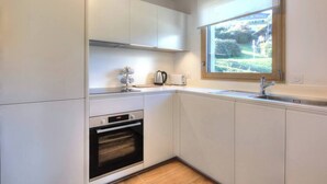 A compact kitchen with white cabinets, a built-in oven, a window with a view of greenery, and a stainless steel sink.