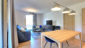 A modern living room with a wooden dining table, blue chairs, a grey sofa, and a TV on a black cabinet.