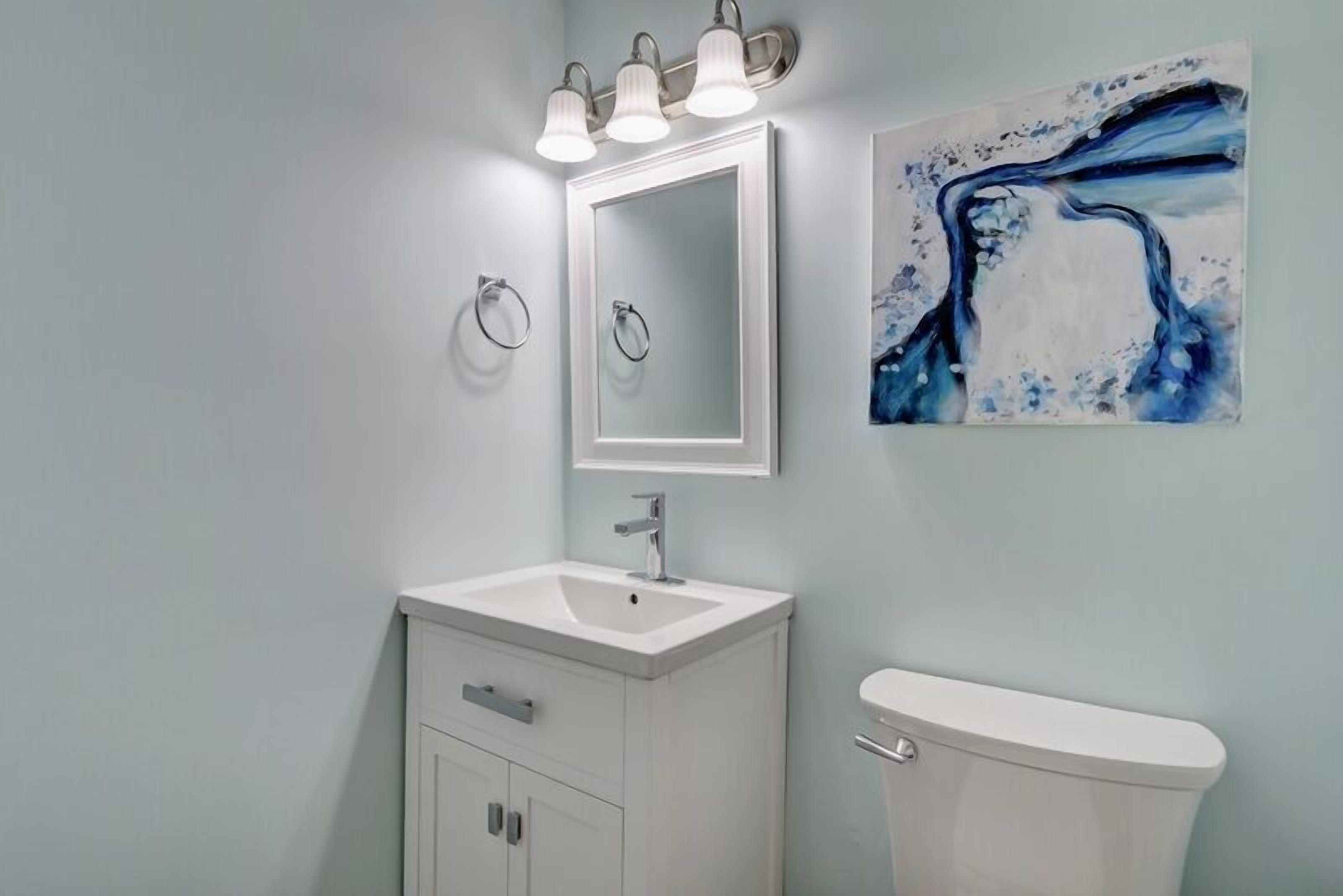 Un baño con un lavamanos blanco, un espejo y un inodoro. Hay un cuadro en la pared y una lámpara sobre el lavamanos.