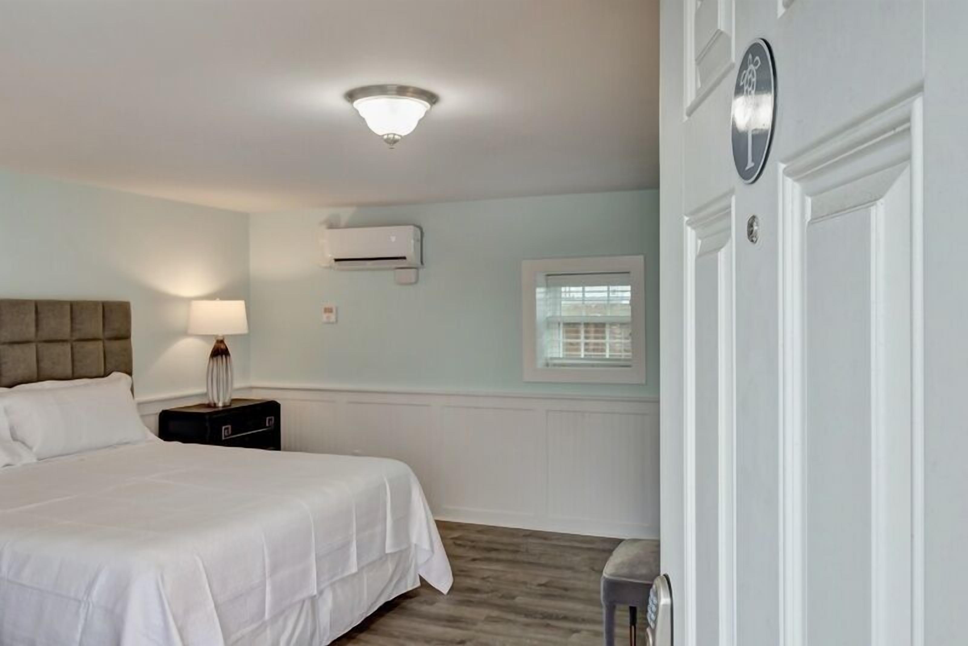 A hotel room with a bed, bedside table, lamp, air conditioning unit, window, and a door with a sign.