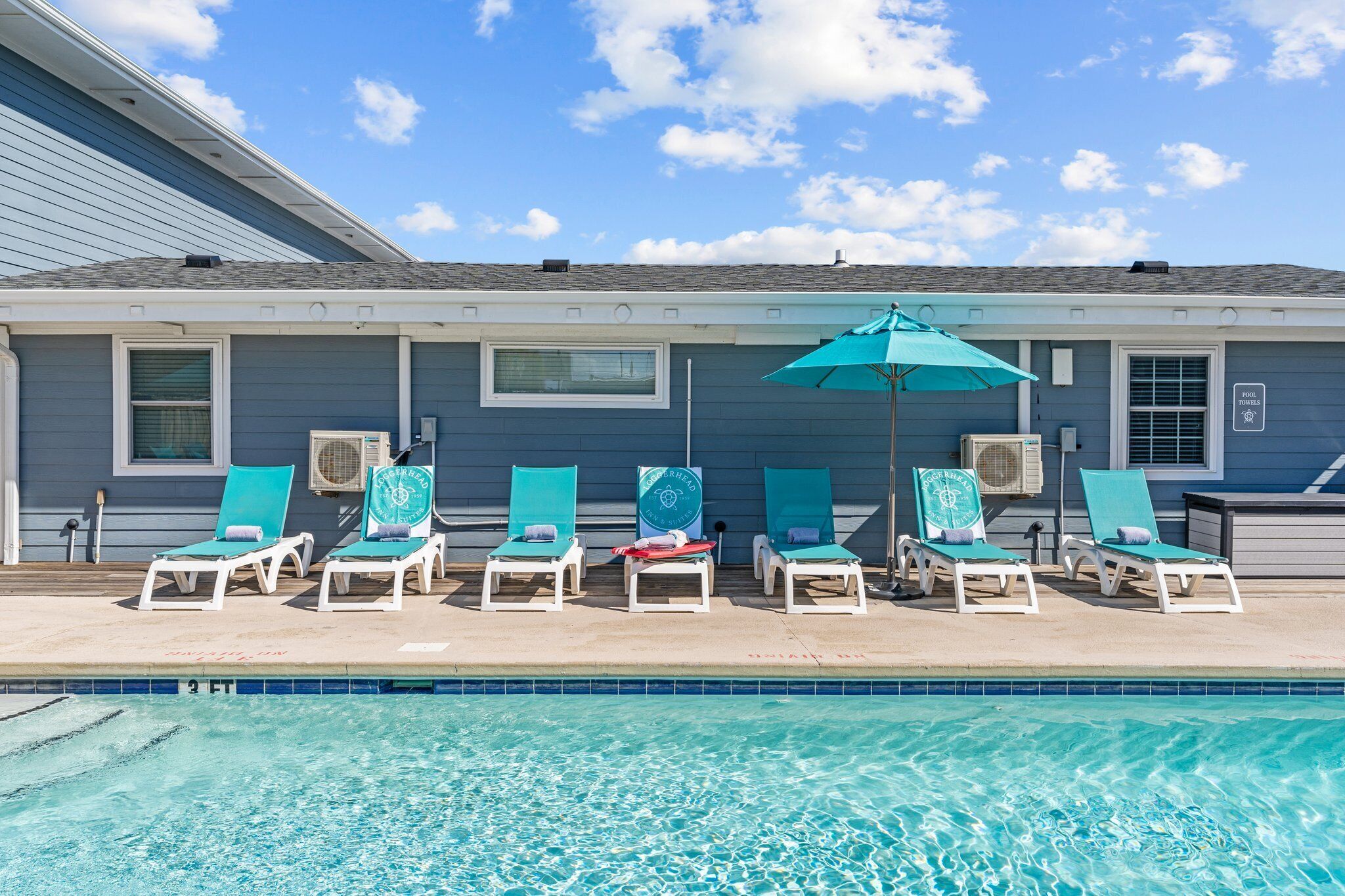A poolside area with lounge chairs and an umbrella, a clear sky, and a building with a blue exterior.