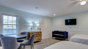 A hotel room with a bed, a desk with two chairs, a dresser, a television, and a window with blinds. - Loggerhead Inn & Suites - Two Queen Suite in hotel in Surf City, North Carolina (Surf City)
