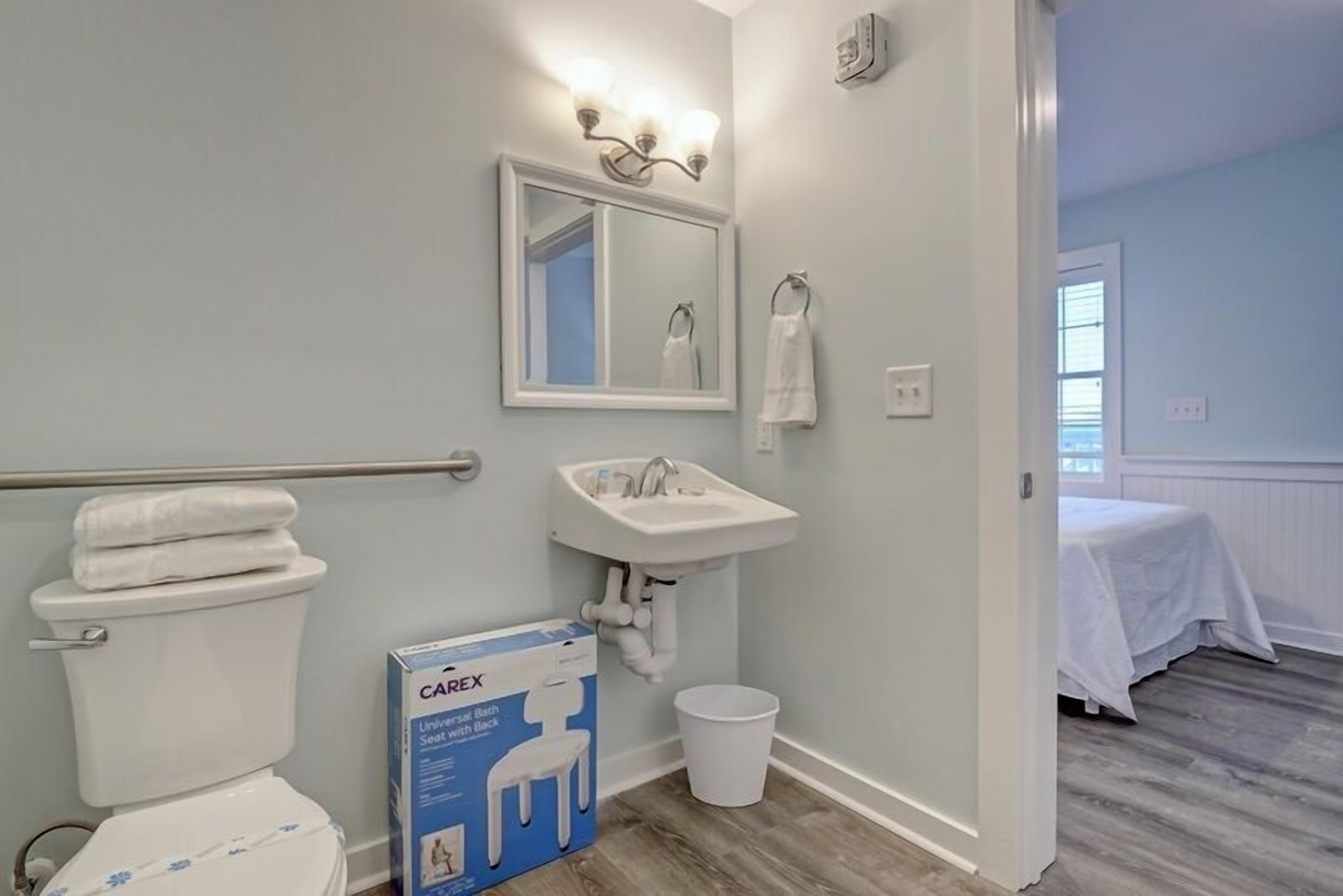 A bathroom with a toilet, sink, and mirror. There is a box labeled "CAREX Universal Bath Seat with Back" on the floor.