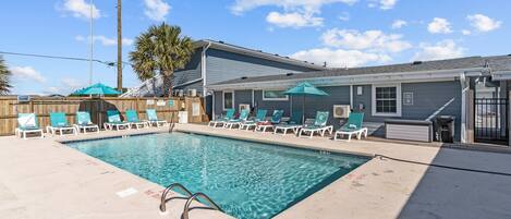 A swimming pool with lounge chairs and umbrellas, surrounded by a wooden fence and a building with a grey exterior.