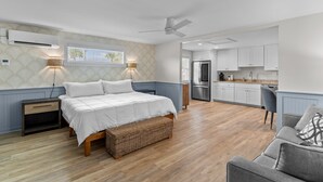 Une chambre à coucher comprenant un lit, des tables de chevet, une commode, un banc en osier, un ventilateur de plafond et une fenêtre avec des stores.