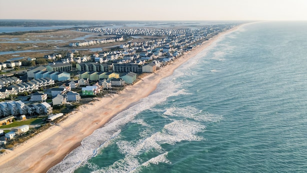 Vue aérienne d’un quartier résidentiel situé sur la côte, avec des maisons alignées le long de la plage.
