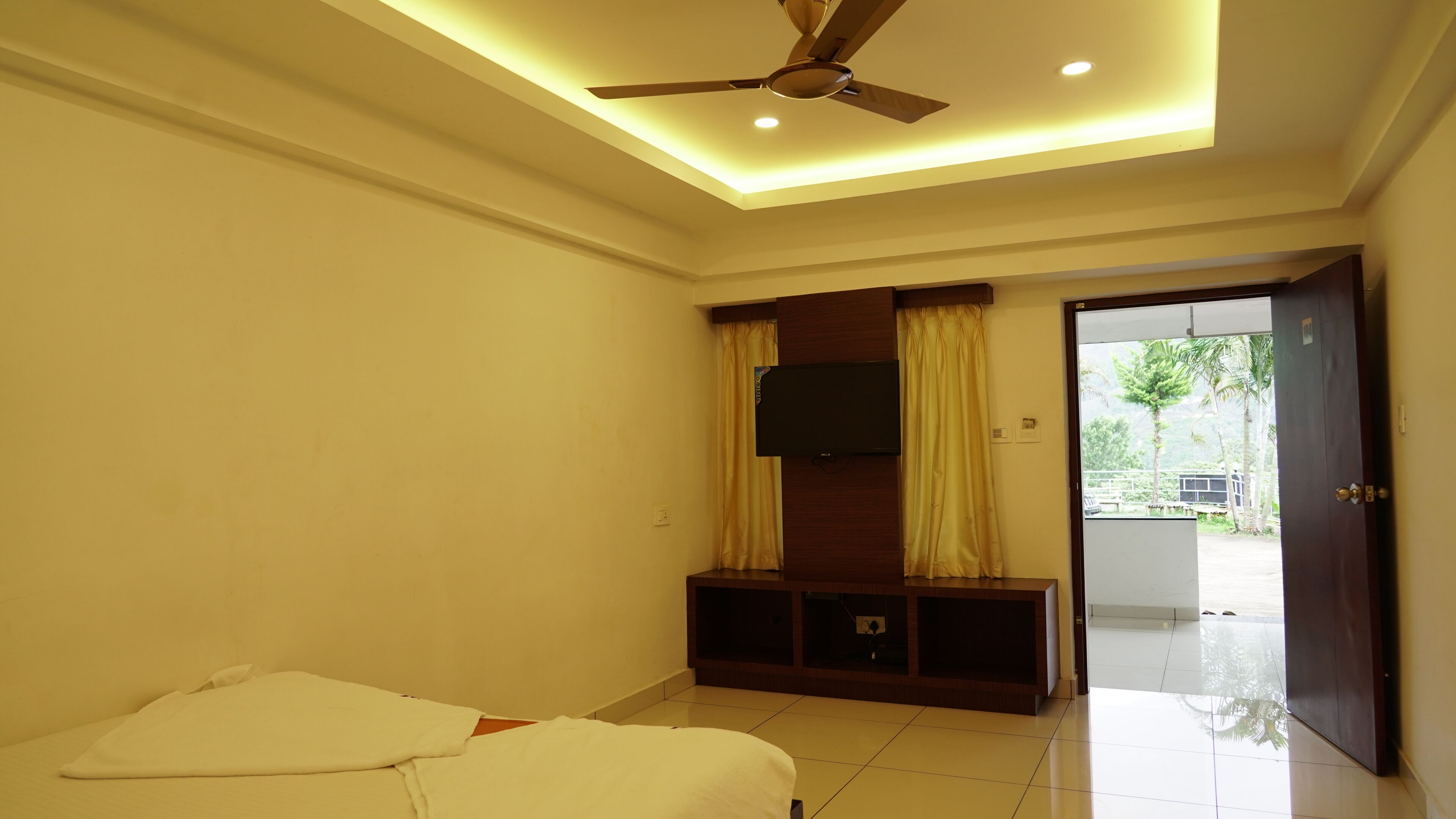 A hotel room with a bed, a TV mounted on a wooden panel, a ceiling fan, and a door leading to a balcony with a view of greenery.