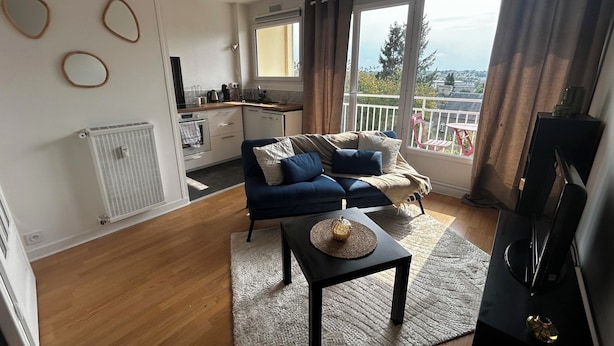 A modern living room with a sofa, a coffee table, and a TV. There is a kitchen area with a window and a balcony with a view. - "Bellevue - 35 m2 - parking - 10 min downtown - Normandy - Saint Lô (Saint-Lô)