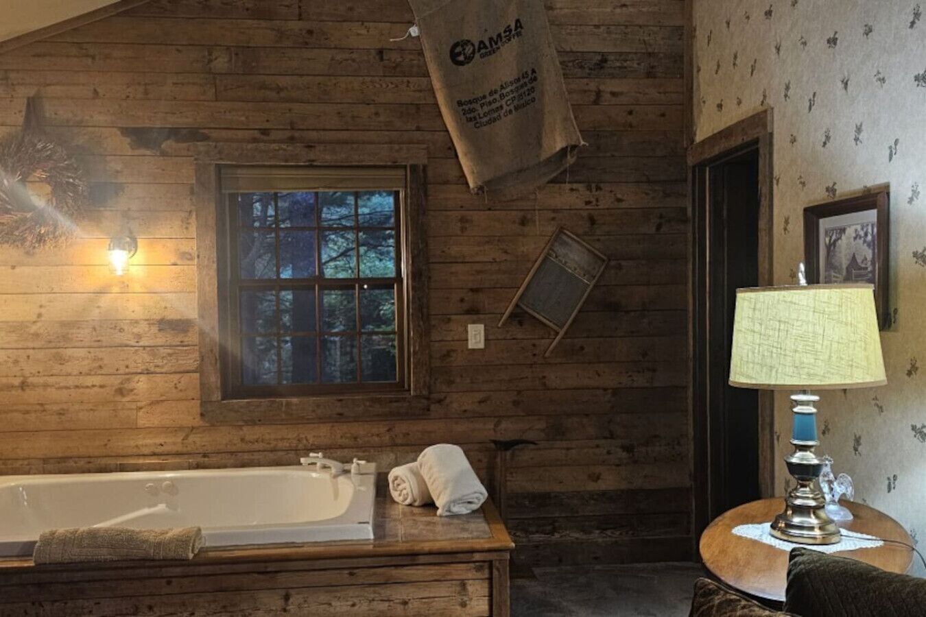 Een rustieke badkamer in chaletstijl met een houten wand, een groot raam, een bad en een klein tafeltje met een lamp.