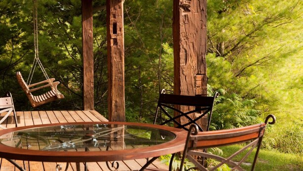 Een houten terras met een tafel met glazen blad en metalen stoelen, omringd door bomen.