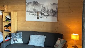 Una sala de estar acogedora con un sofá gris, estanterías de madera, un cuadro colgado en la pared que muestra una escena de montaña nevada y una lámpara de mesa.