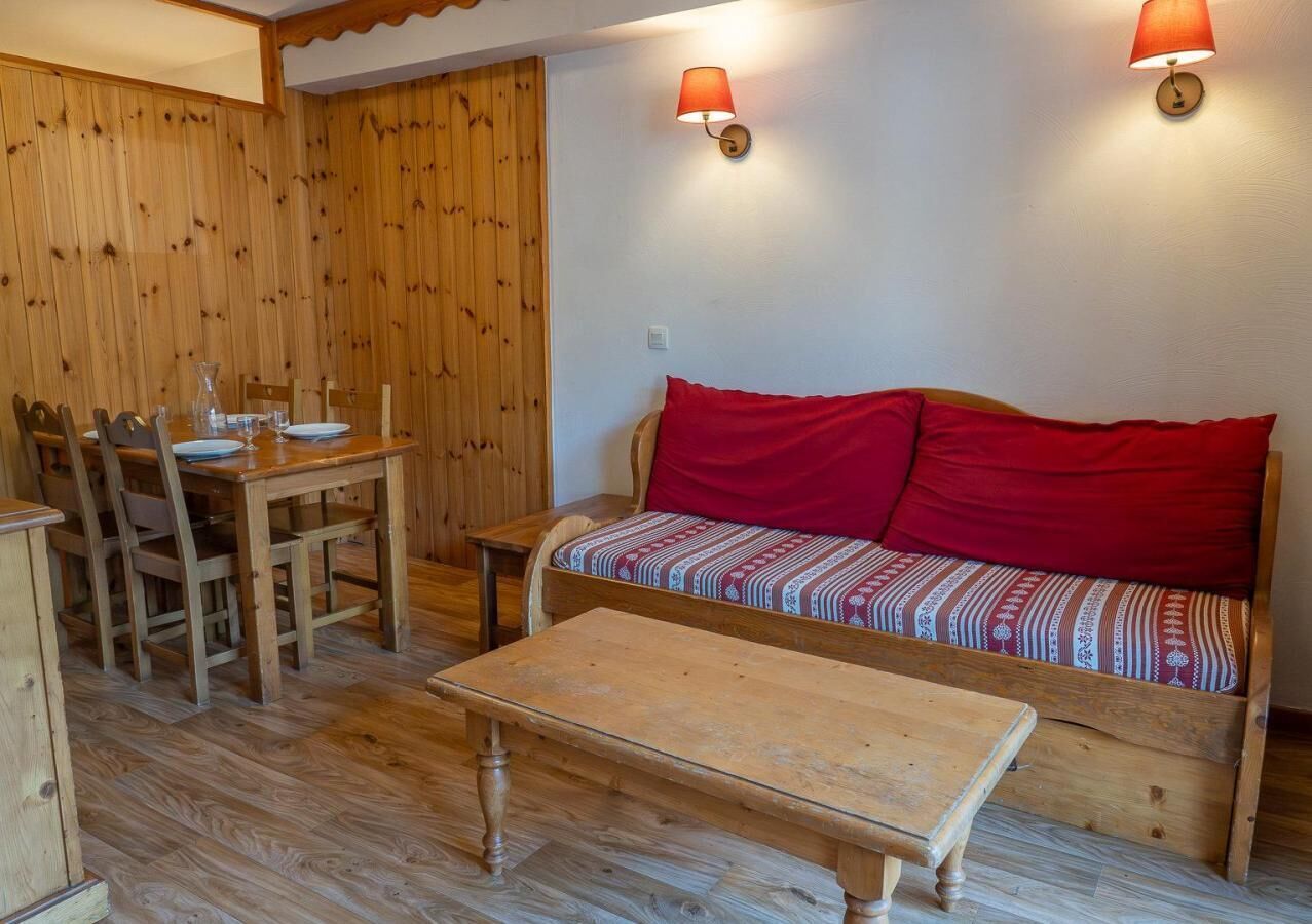 Ein gemütliches Zimmer mit einem Holzesstisch, Stühlen, einer Couch mit roten Kissen und zwei Wandlampen.
