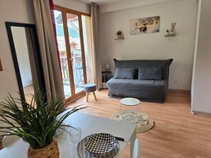 Un salon moderne avec un canapé gris, une table à manger agrémentée d’une plante, et une vue sur une montagne par de grandes fenêtres.