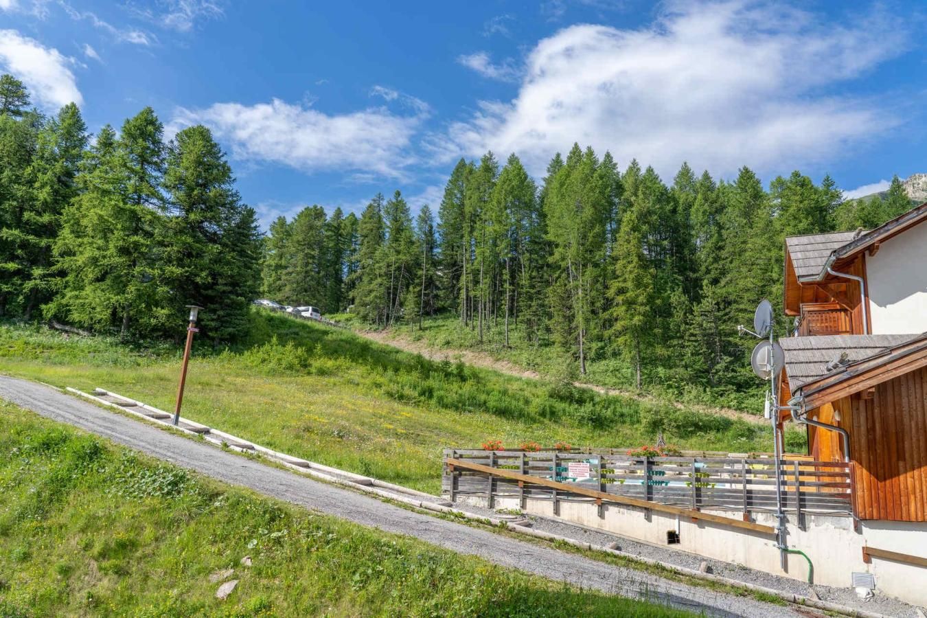 A rural setting with a wooden cabin, a gravel road, and a forested hillside.