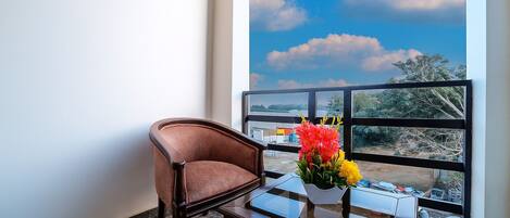 A room with a view of a clear sky and clouds, a window, a chair, and a table with a vase of flowers.